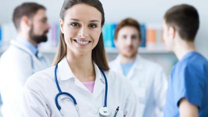 Vorsorgecheck - Freundliche Ärztin lächelt in die Kamera mit Arztkollegen im Hintergrund